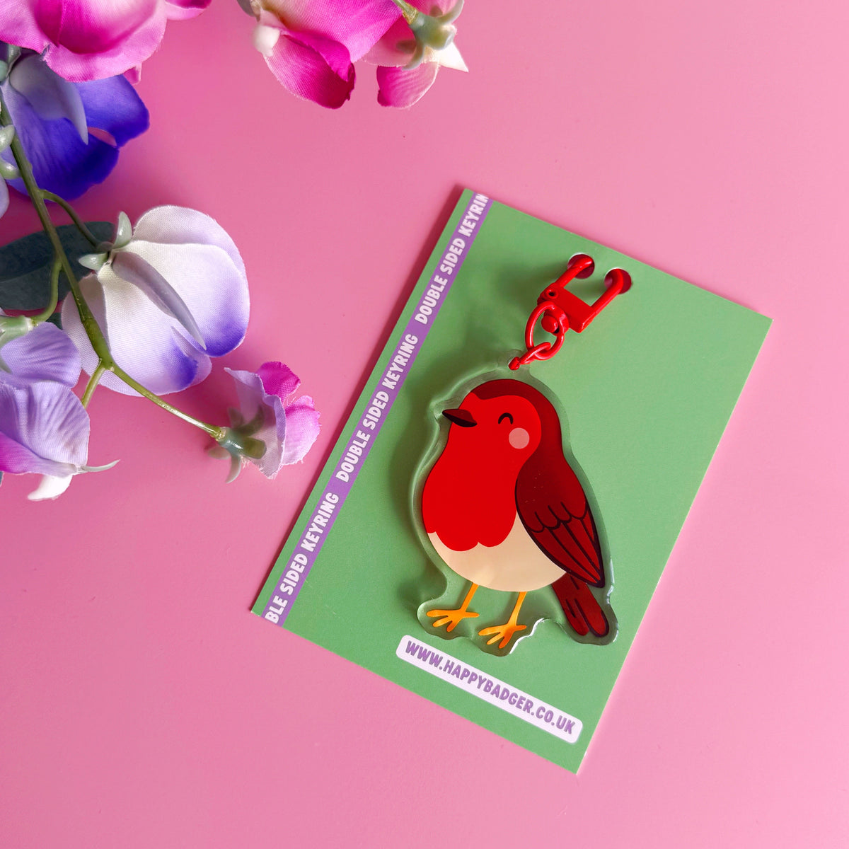 A colourful robin keychain or bag charm attached to a green backing card, photographed on a pink background with decorative flowers in the top left corner.
