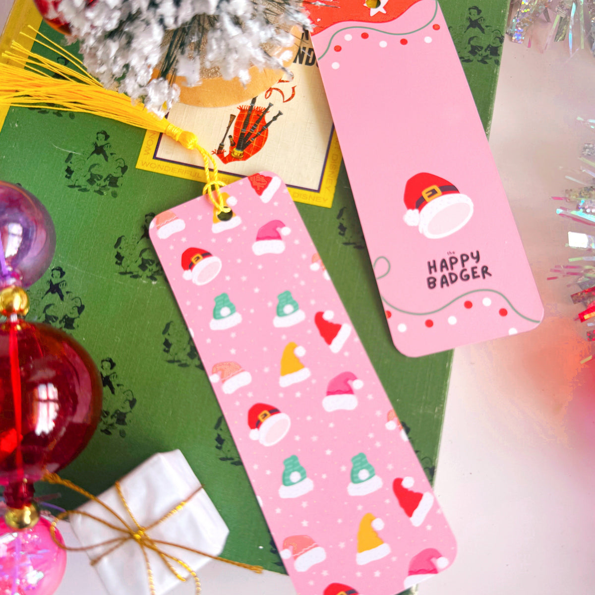 An image showing the front and reverse side of a Christmas bookmark - that has multicoloured Santa and eElf hats on the front, and a festive pattern to the back with the Happy Badger branding logo.