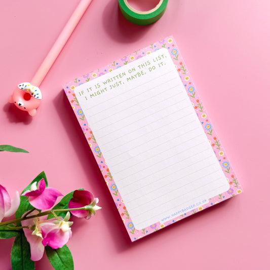Notepad with decorative border on a pink background with pen, tape, and flowers.