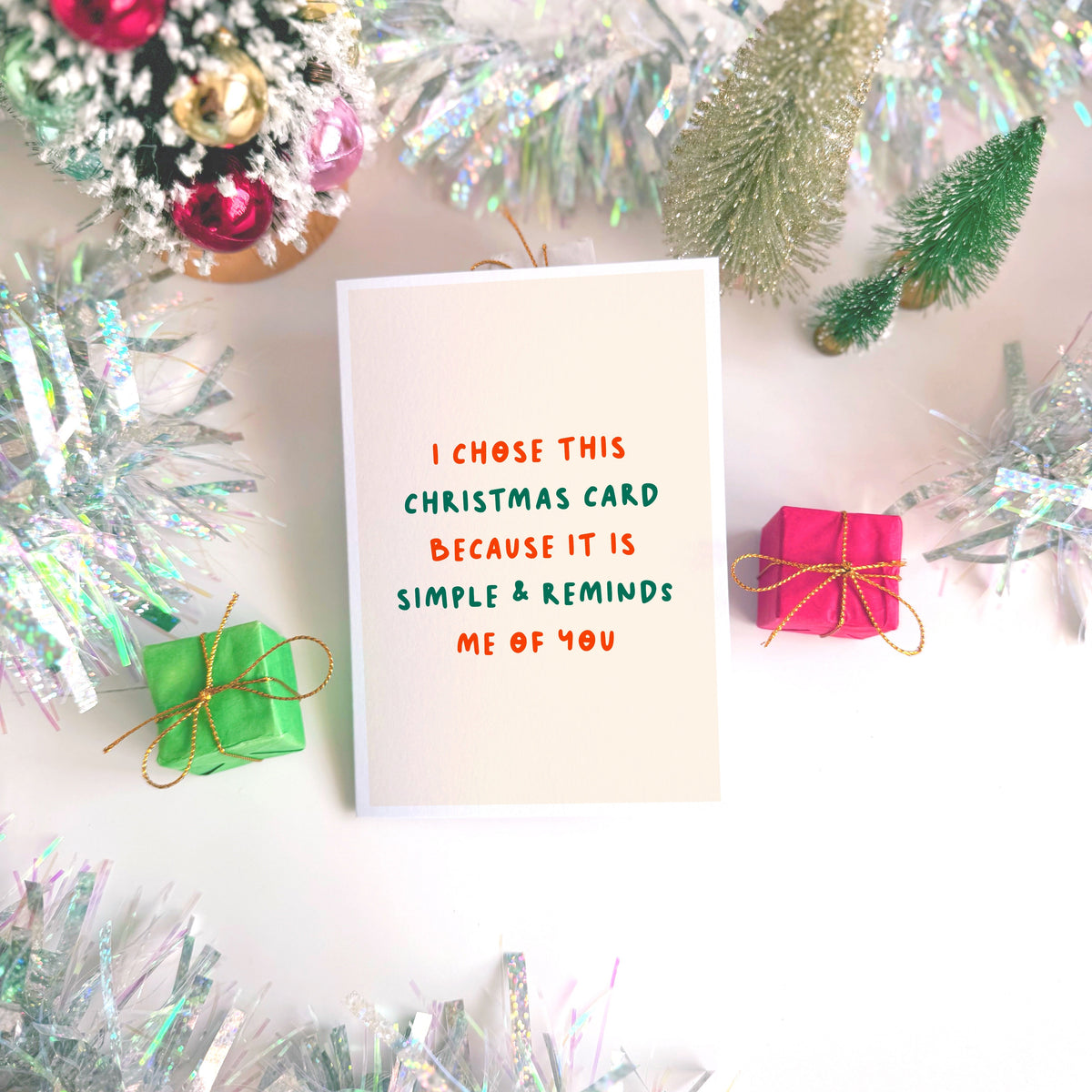 A minimalist Christmas greeting card with a warm cream background and red and green text reading 'I CHOSE THIS CHRISTMAS CARD BECAUSE IT IS SIMPLE & REMINDS ME OF YOU'.