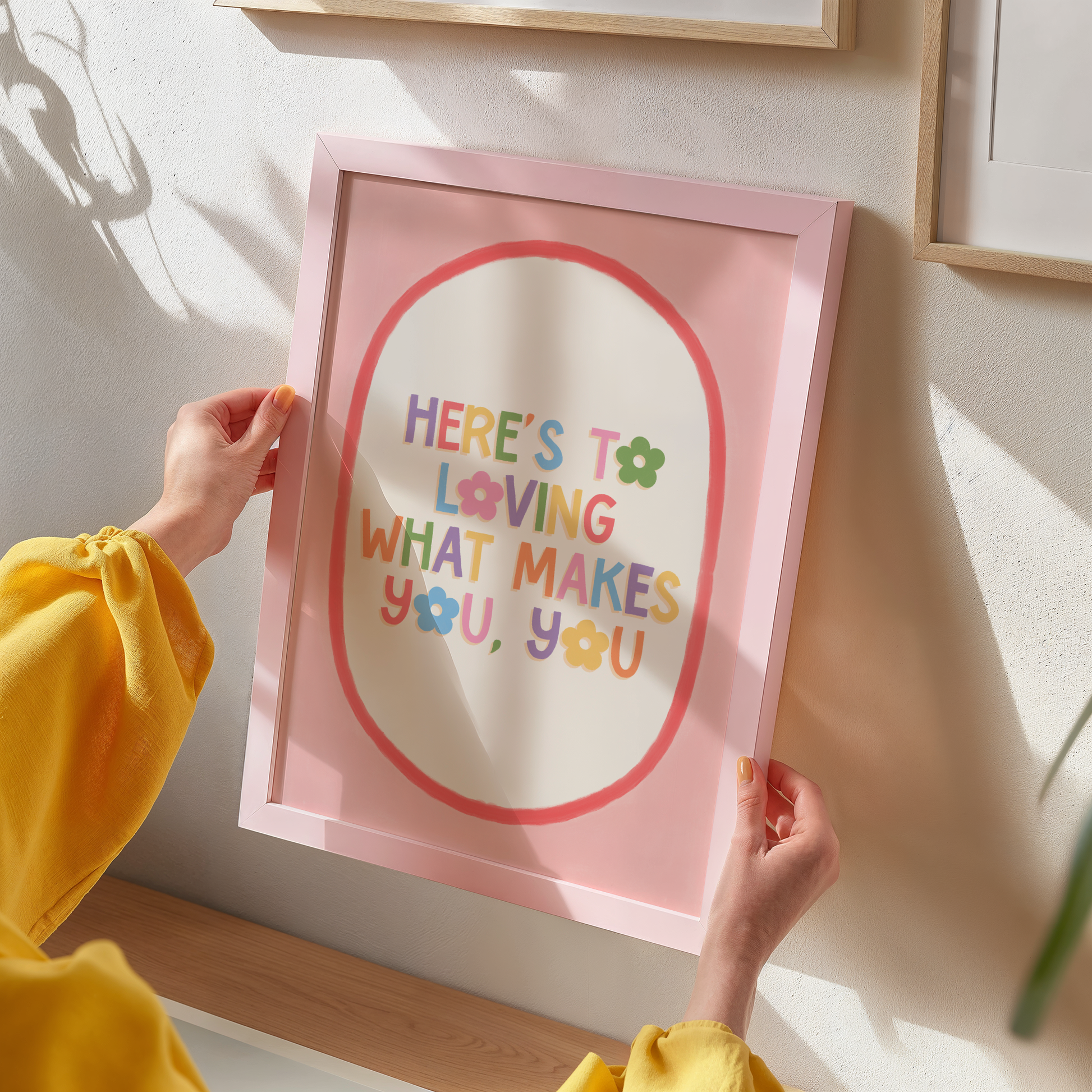 A positive quote print that reads 'here's to loving what makes you, you' in multicoloured text with a pink and red oval shaped border.