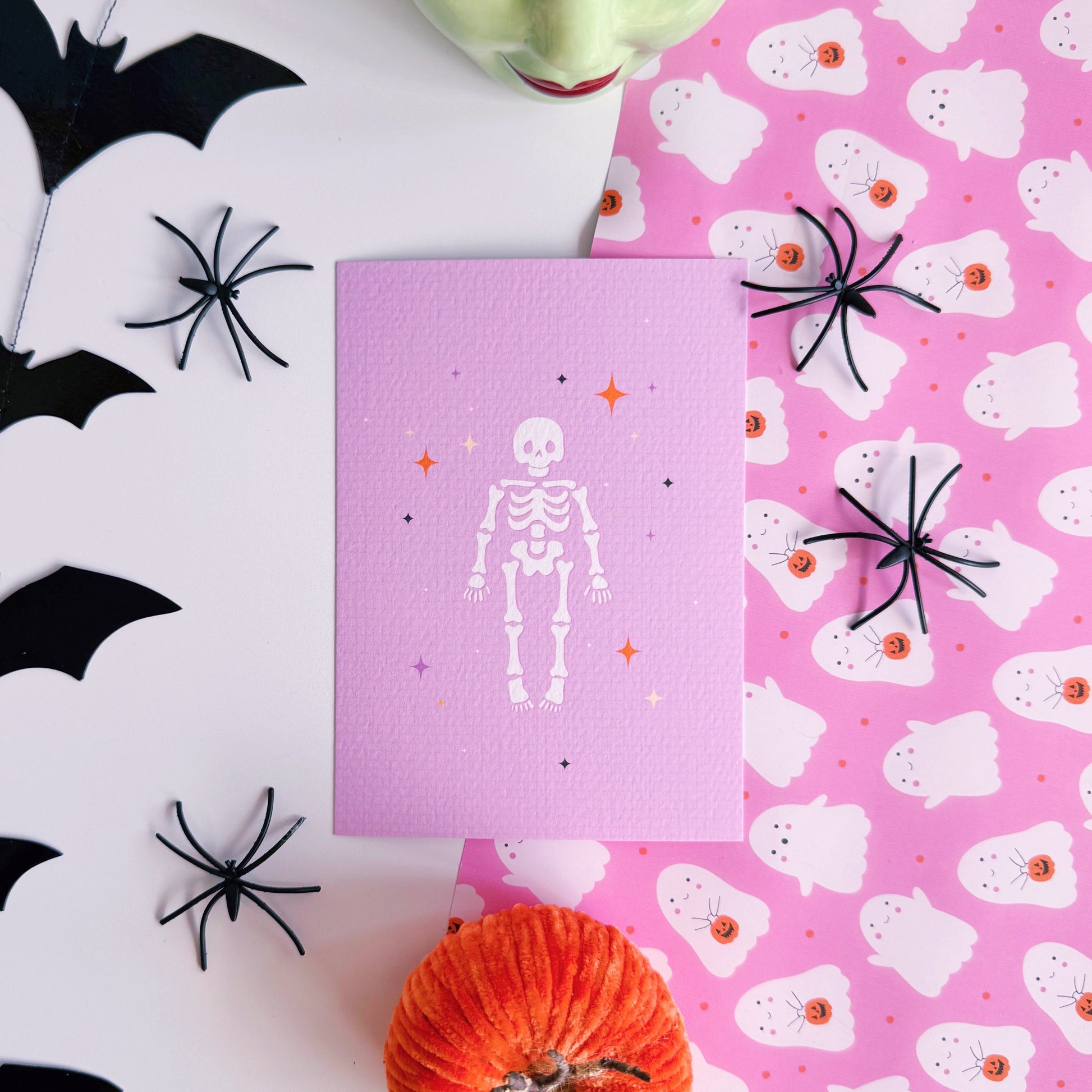 A purple Halloween postcard with an illustration of a skeleton on the front, surrounded by little multi-coloured stars.
