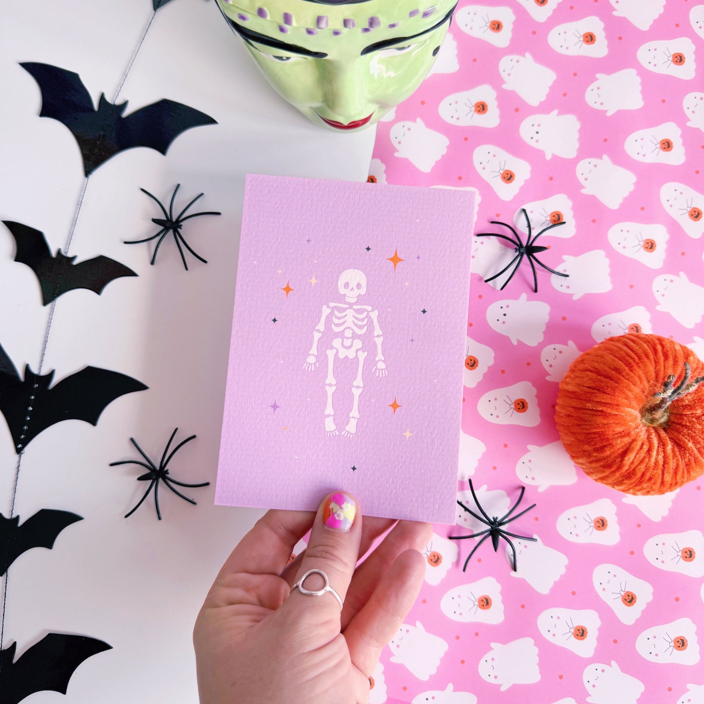 A purple Halloween postcard with an illustration of a skeleton on the front, surrounded by little multi-coloured stars.