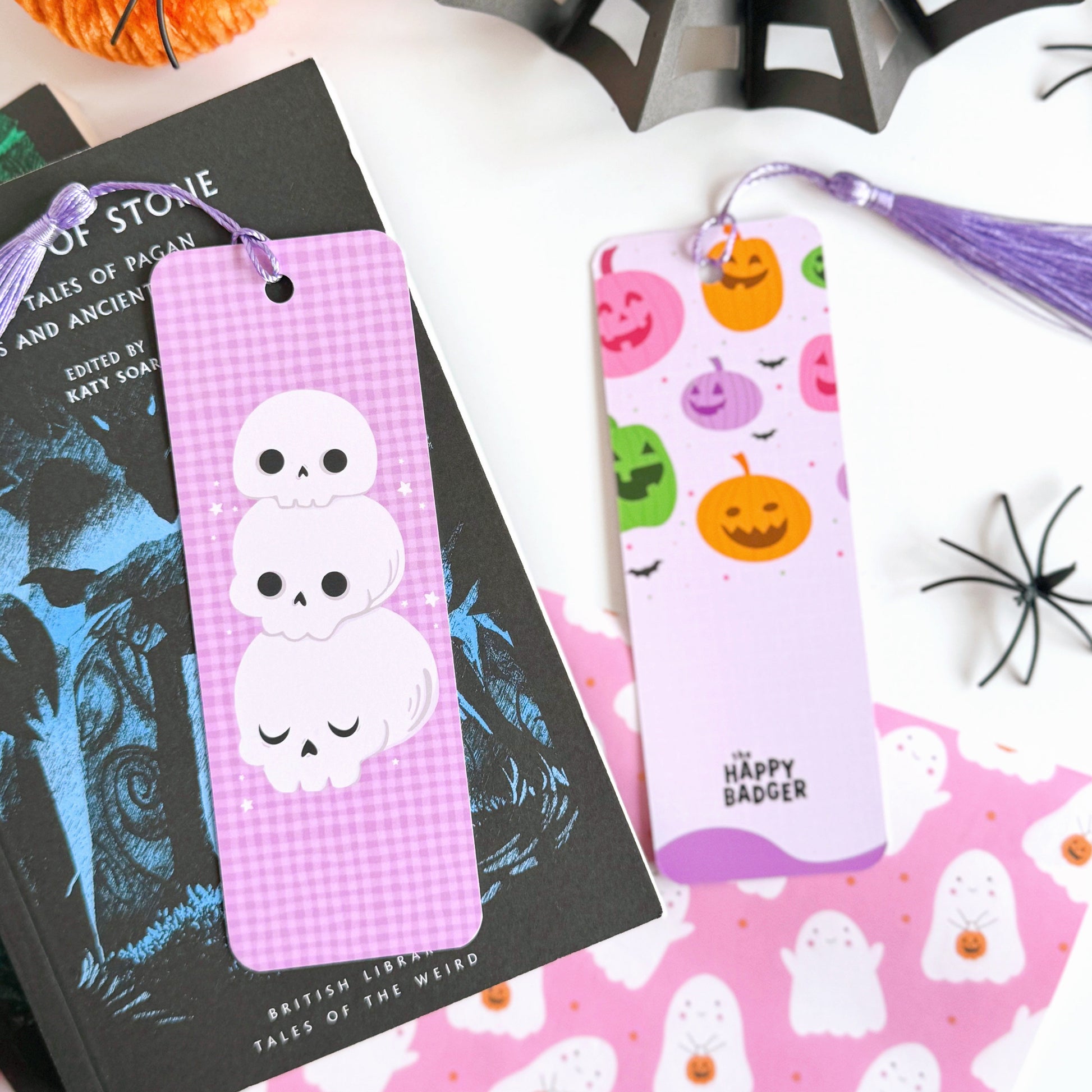 A purple gingham patterned bookmark, with a skull illustration on the front. On the back are pumpkins and the brand logo. The bookmark has a purple tassel attached. 