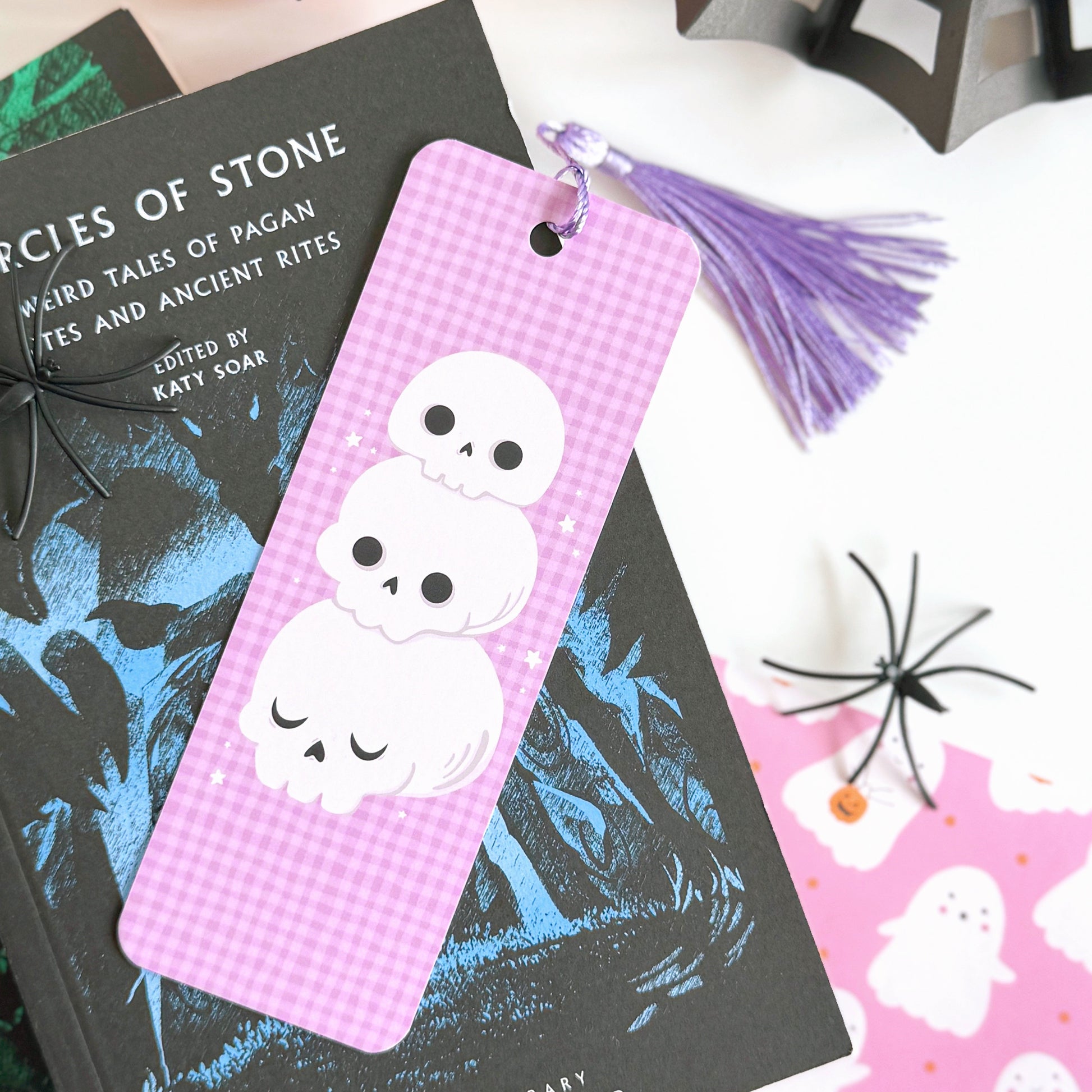 A purple gingham patterned bookmark, with a skull illustration on the front. On the back are pumpkins and the brand logo. The bookmark has a purple tassel attached. 