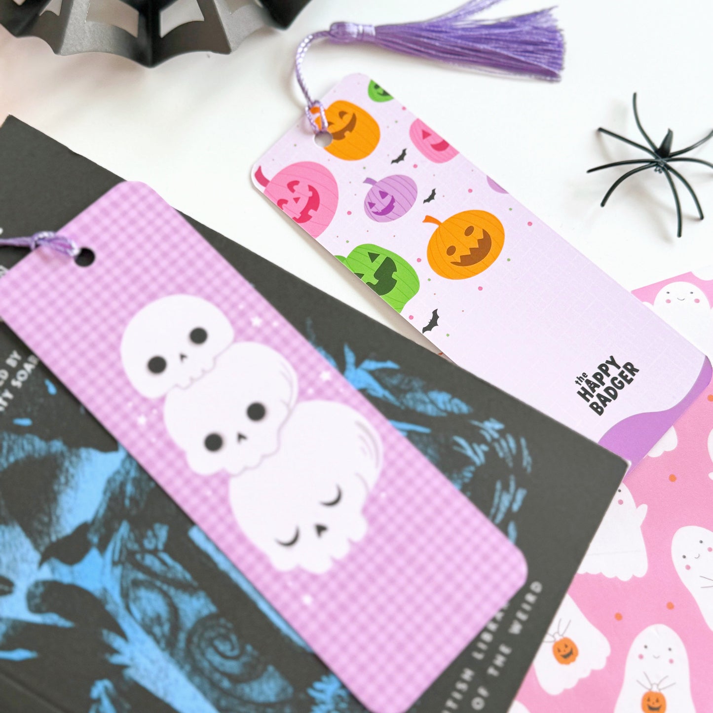 A purple gingham patterned bookmark, with a skull illustration on the front. On the back are pumpkins and the brand logo. The bookmark has a purple tassel attached. 