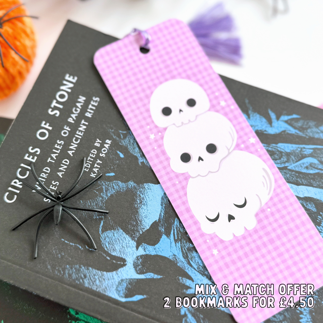 A purple gingham patterned bookmark, with a skull illustration on the front. On the back are pumpkins and the brand logo. The bookmark has a purple tassel attached. 