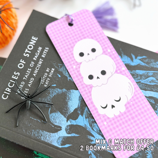 A purple gingham patterned bookmark, with a skull illustration on the front. On the back are pumpkins and the brand logo. The bookmark has a purple tassel attached. 