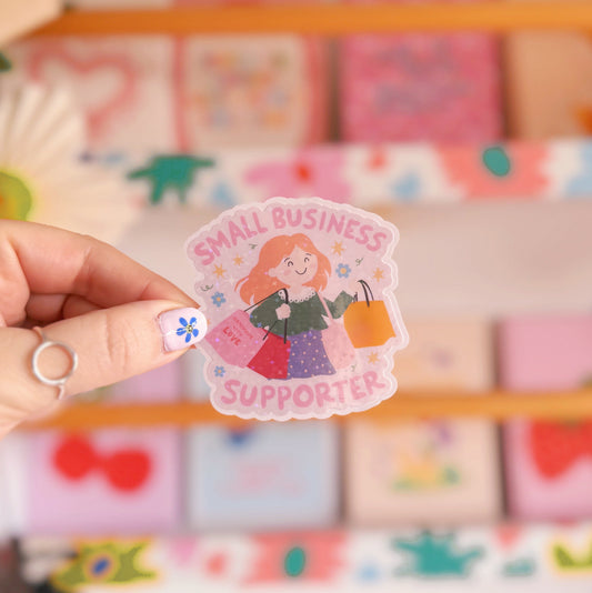 A holographic sticker showing a woman with red hair carrying lots of shopping bags and the words 'small business supporter' around the illustration.