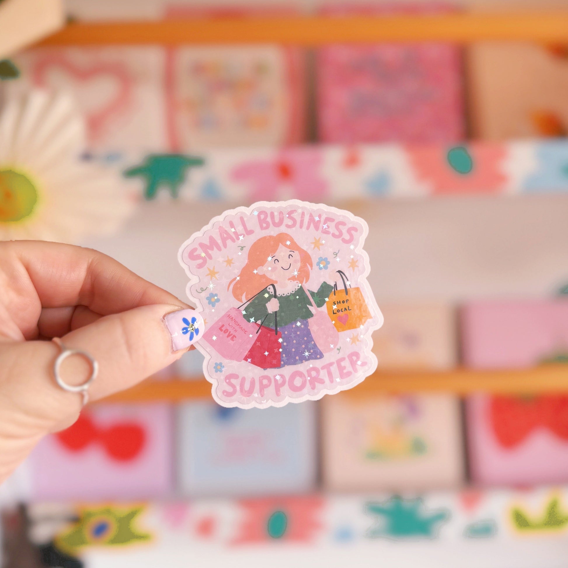A holographic sticker showing a woman with red hair carrying lots of shopping bags and the words 'small business supporter' around the illustration.