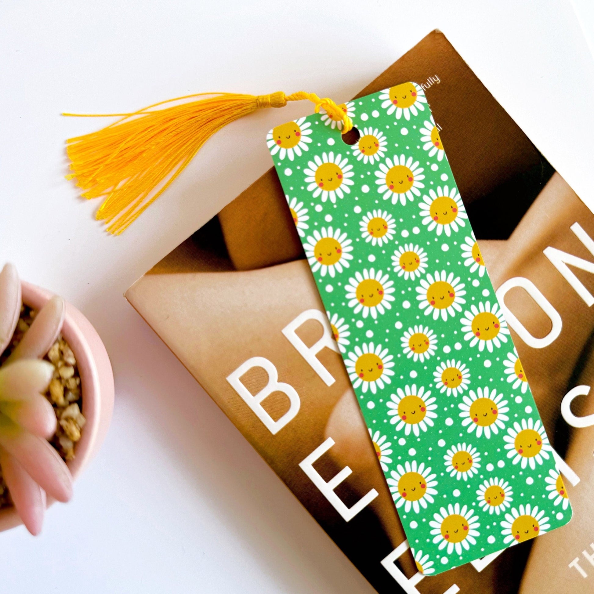 A picture of a green bookmark, with a coordinating yellow tassel, with a repeating pattern of white and yellow smiling flowers on it.