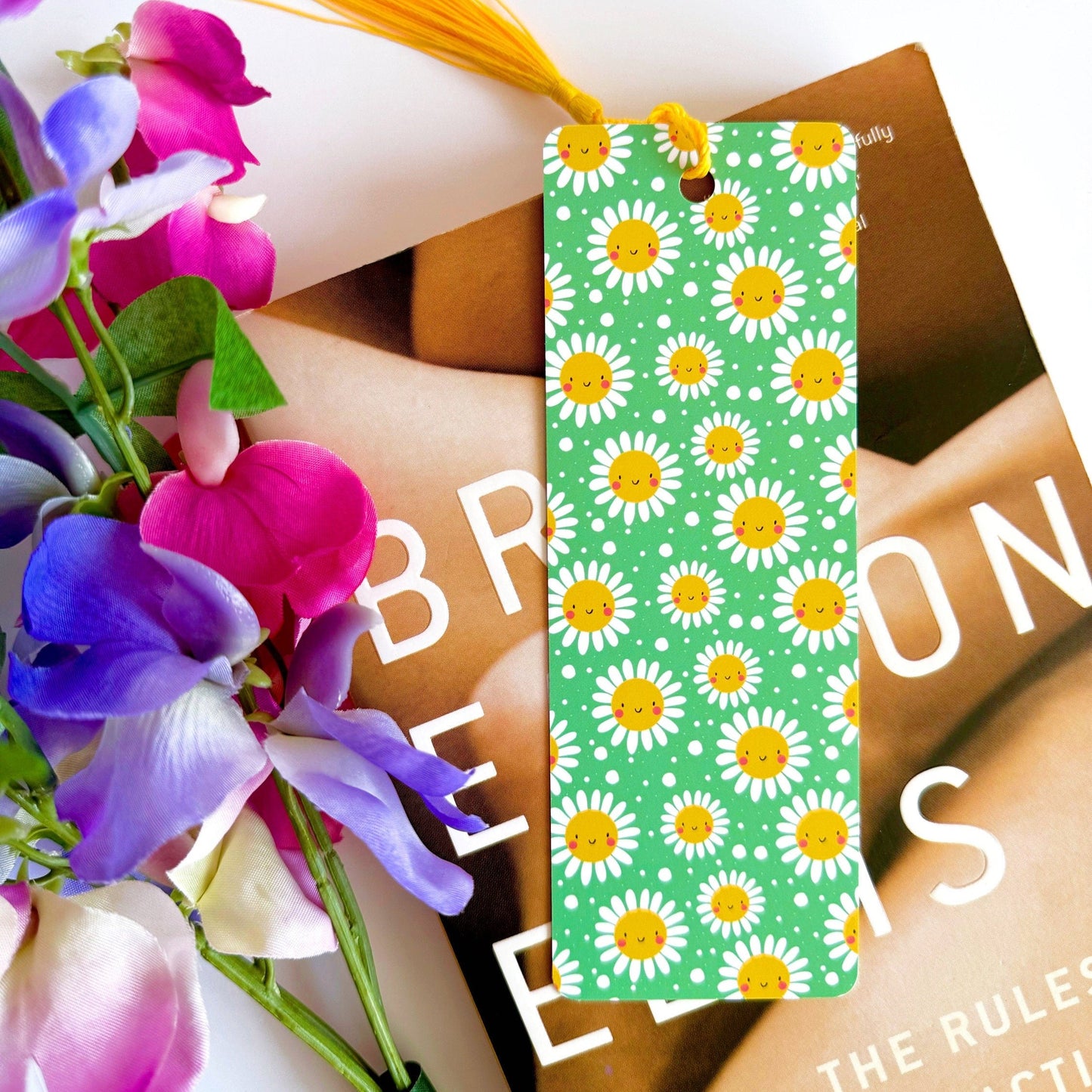 A picture of a green bookmark, with a coordinating yellow tassel, with a repeating pattern of white and yellow smiling flowers on it.