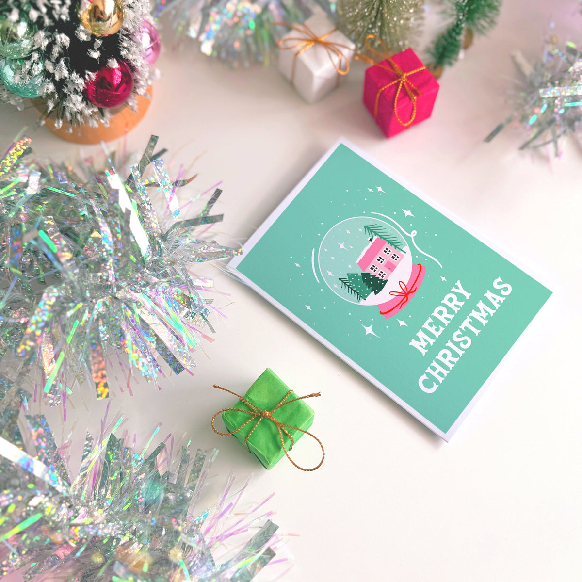 A festive Christmas greeting card with a mint green background, featuring an illustration of a snow globe house and the words 'MERRY CHRISTMAS' in white. The card is styled in a holiday flat-lay arrangement with miniature Christmas trees, silver tinsel, small wrapped gift boxes, and a white surface.