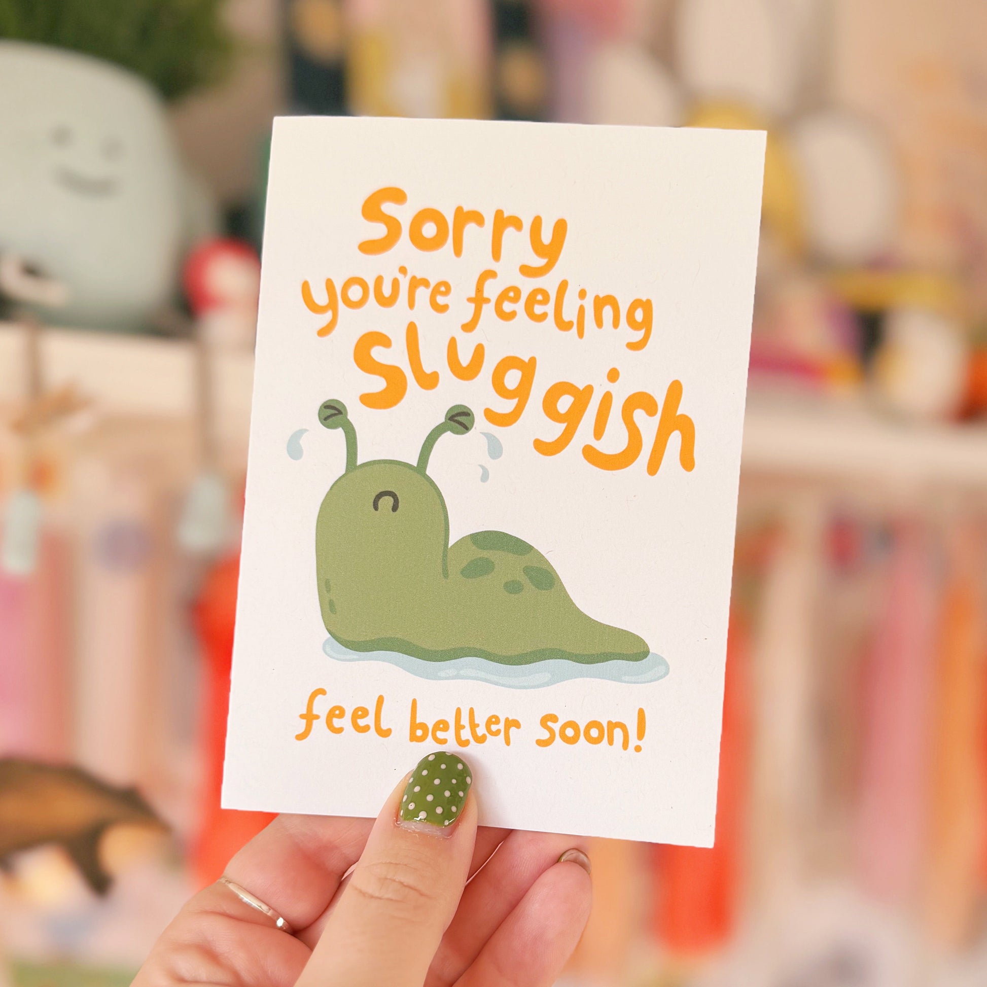 Hand holding a card with a cartoon slug and text 'Sorry you're feeling sluggish feel better soon!' in a blurred indoor setting.
