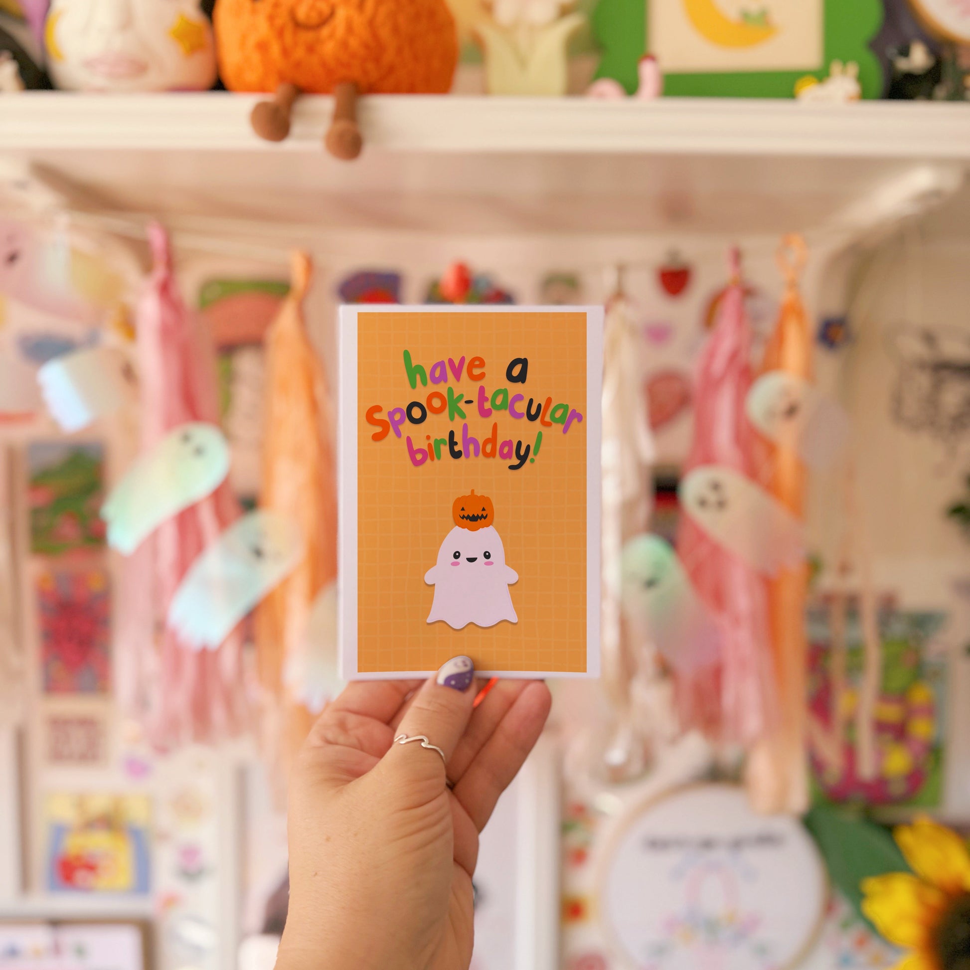 Hand holding a 'have a spooktacular birthday!' card in front of a colorful shelf with decorations.
