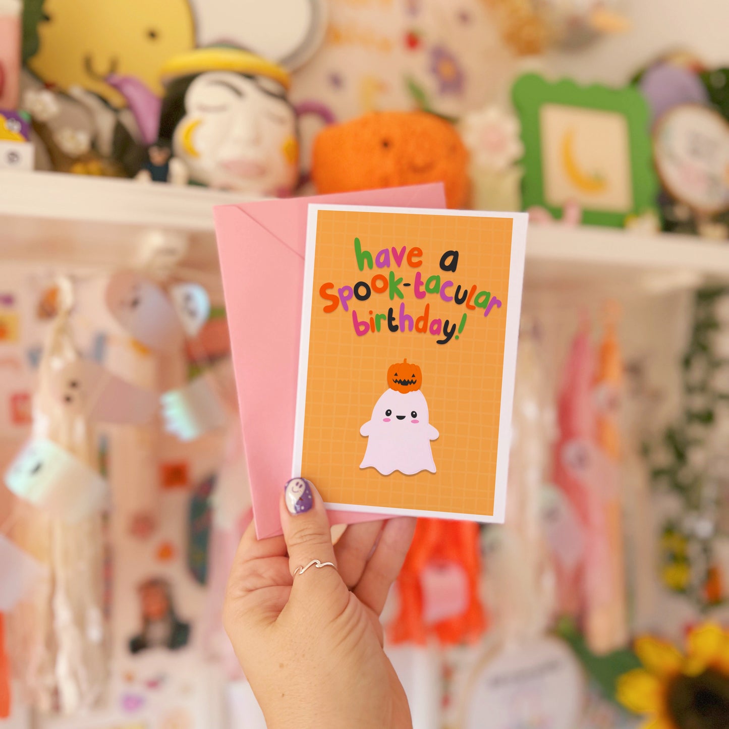 Hand holding a Halloween-themed birthday card with a ghost and pumpkin design in front of a colorful background.