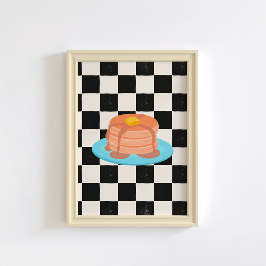 A framed art print featuring a black and white checkered pattern with a colourful illustration of pancakes on a blue plate.