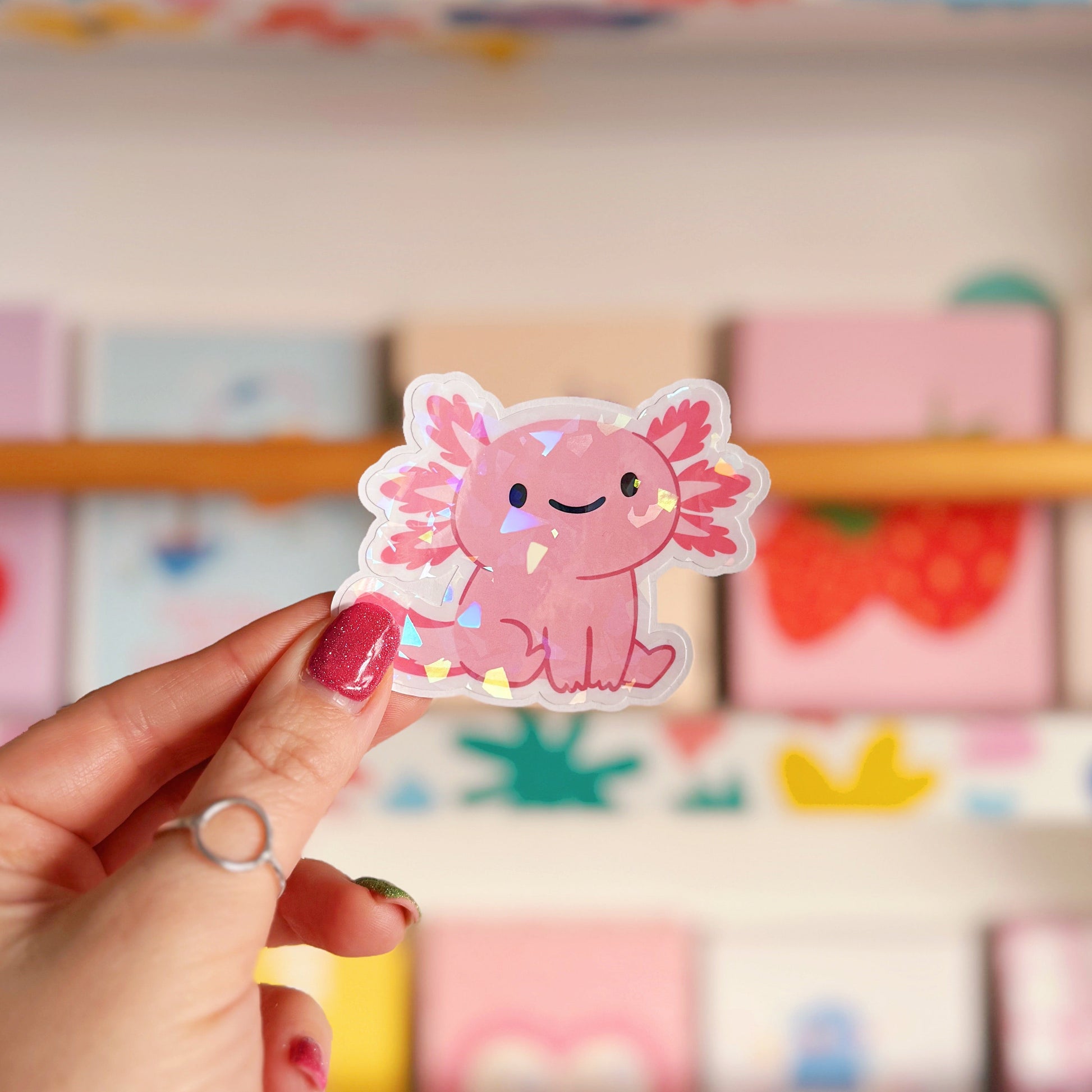 Hand holding a pink axolotl sticker in front of a colourful background