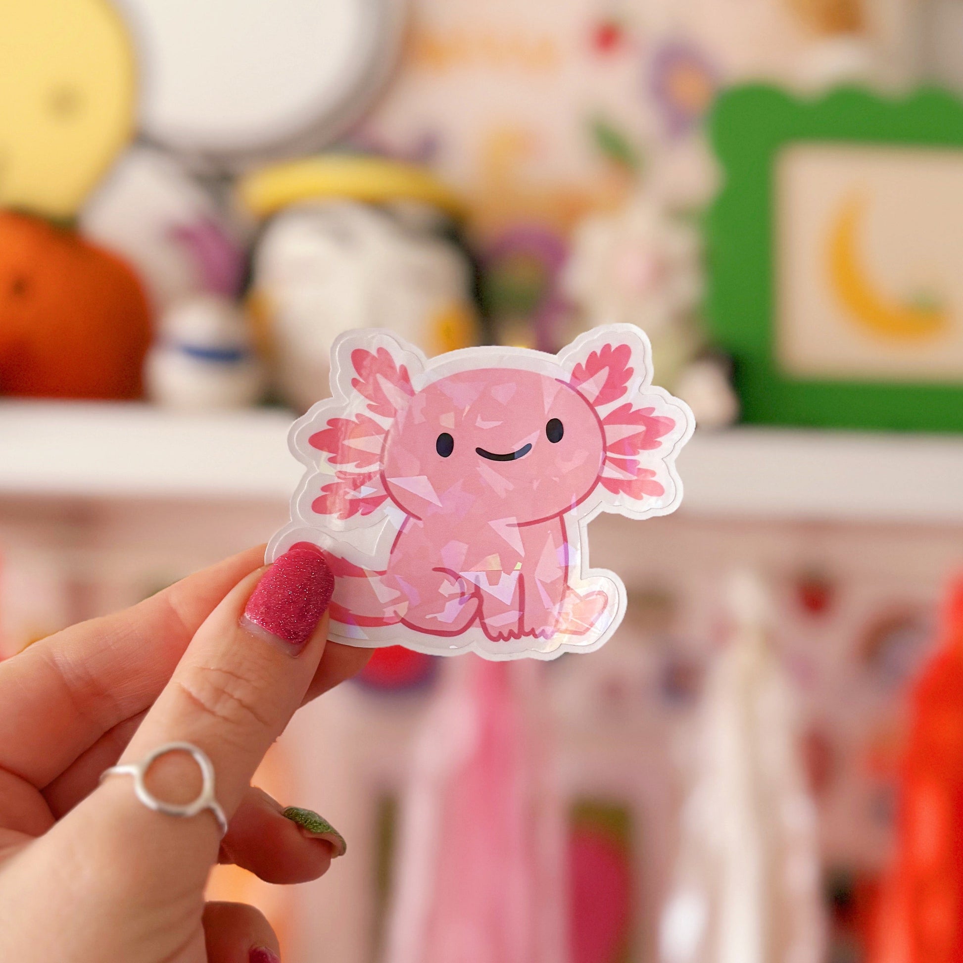 Hand holding a pink axolotl sticker in front of a colourful background