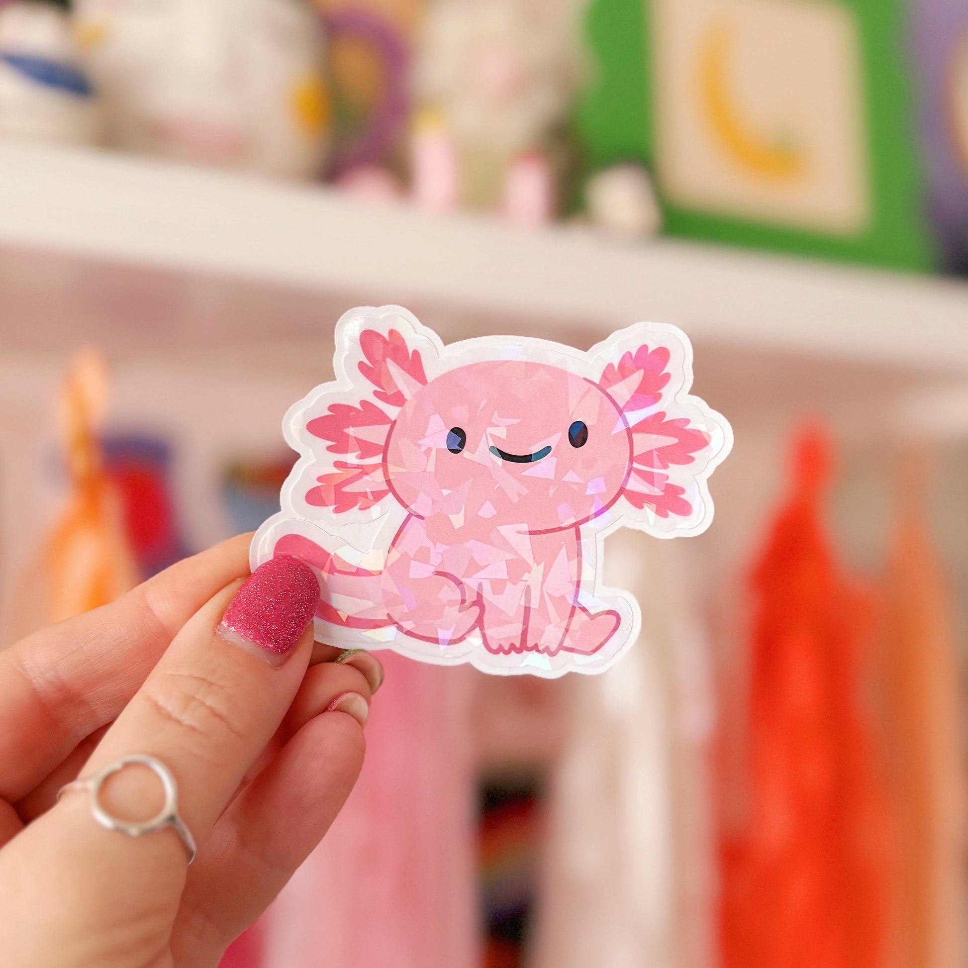 Hand holding a pink axolotl sticker in front of a colourful background