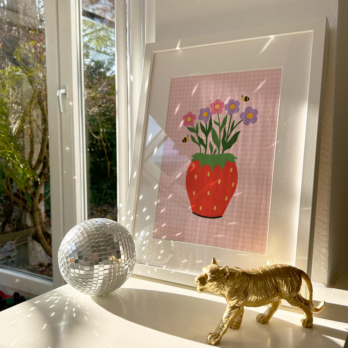 Framed artwork of a strawberry with flowers on a windowsill with a disco ball and gold cat figurine.