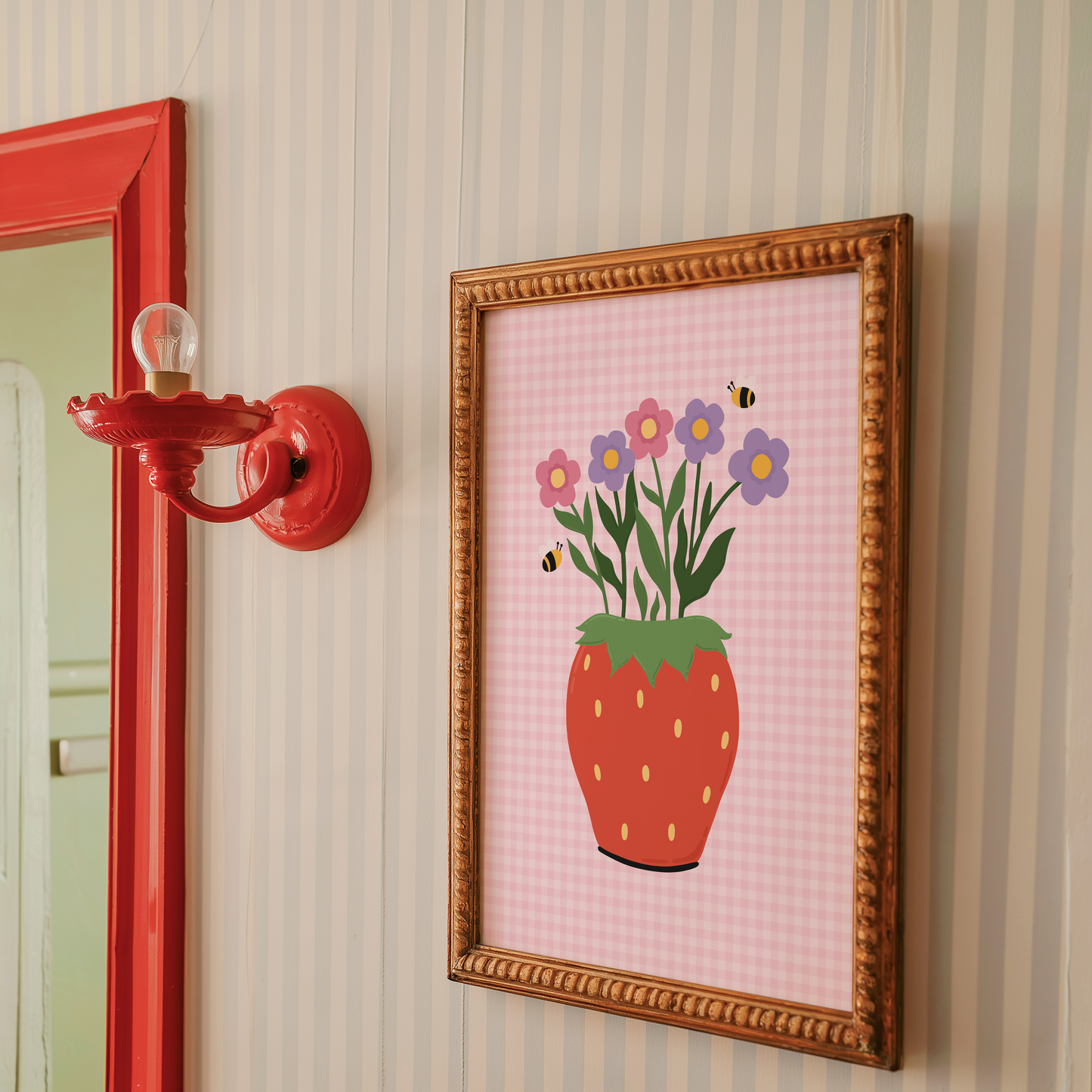 Framed artwork of a strawberry with flowers on a striped wall next to a red mirror.