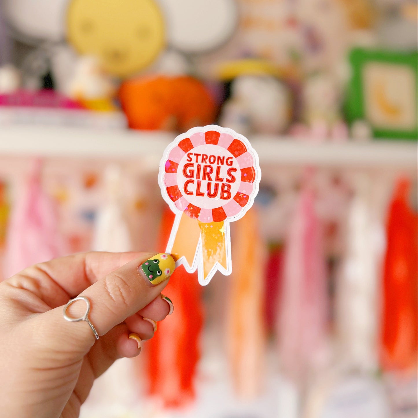 A holographic sticker, with the words 'strong girls club' written in the centre of a rosette award, in pink red and yellow colours.