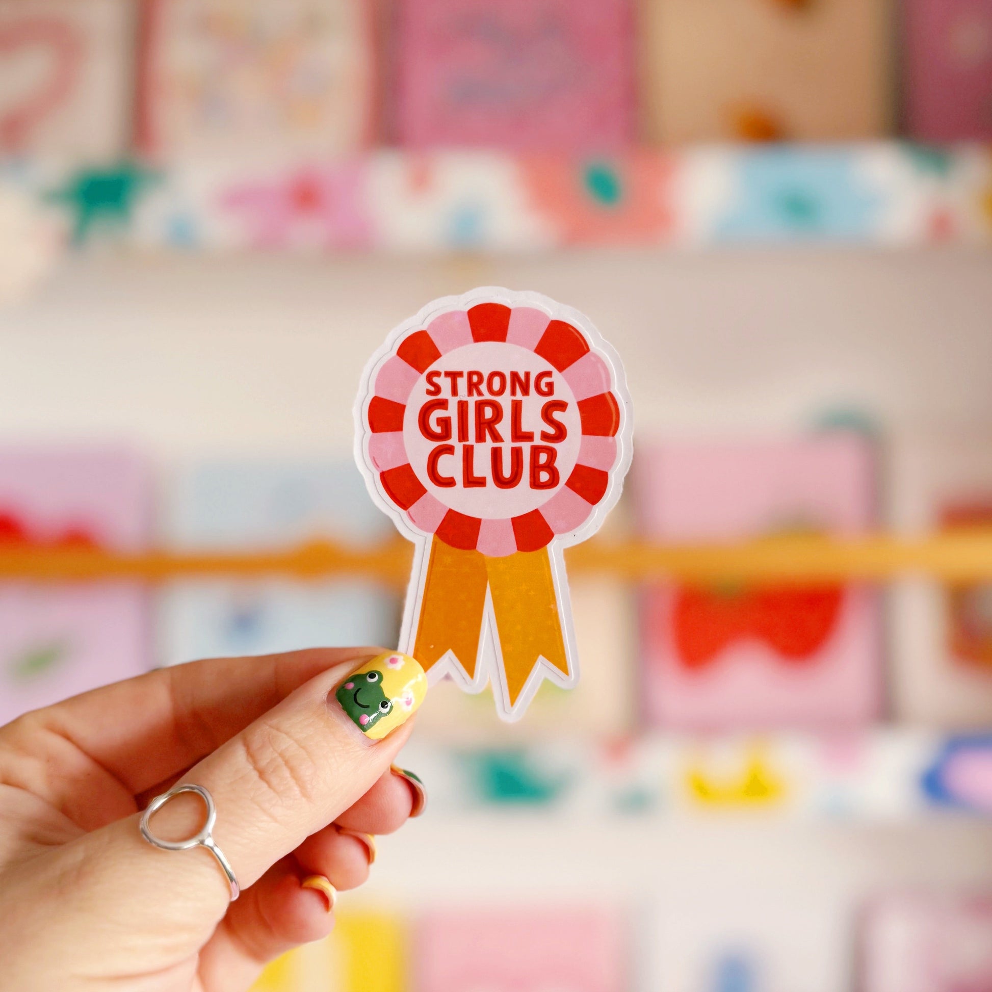 A holographic sticker, with the words 'strong girls club' written in the centre of a rosette award, in pink red and yellow colours.