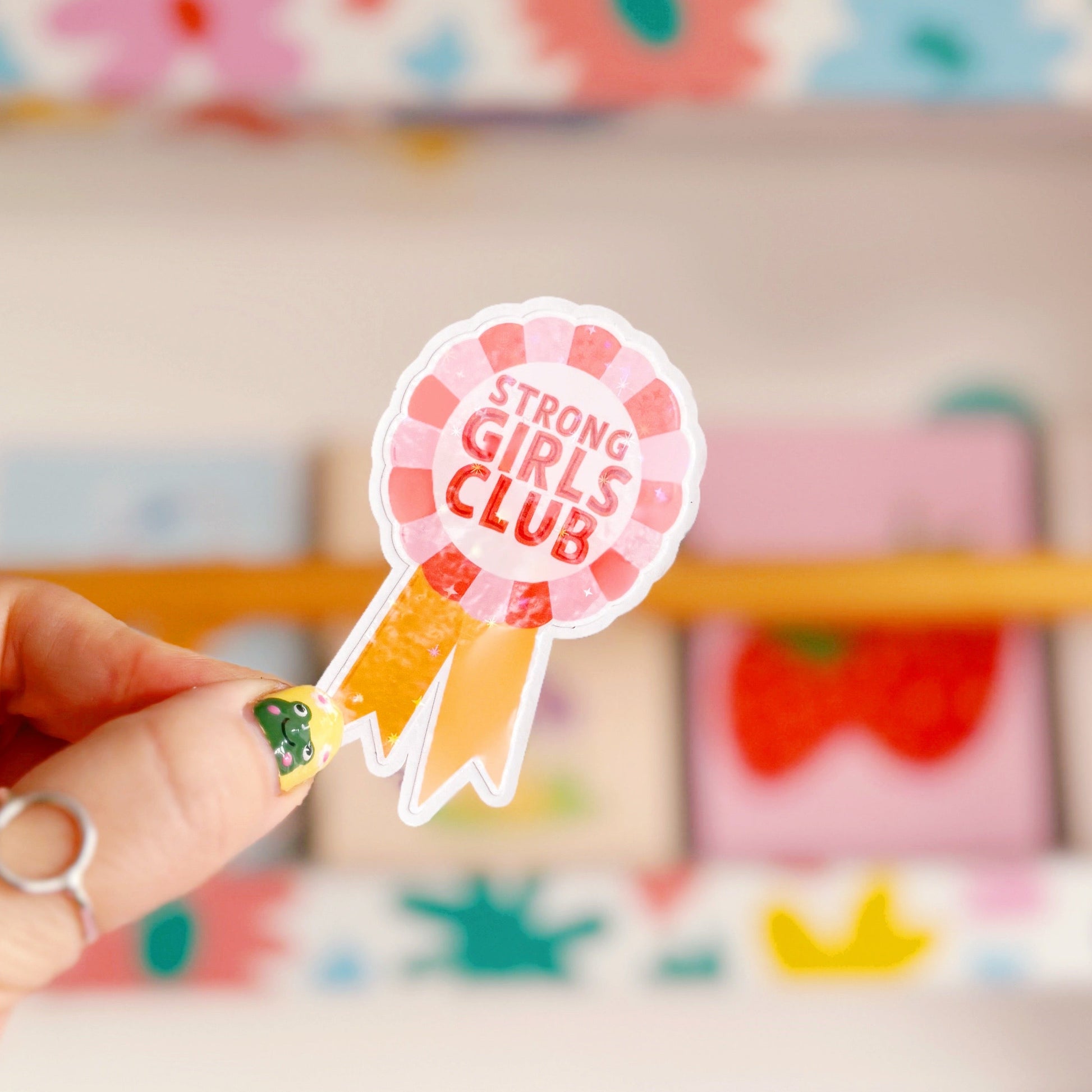 A holographic sticker, with the words 'strong girls club' written in the centre of a rosette award, in pink red and yellow colours.