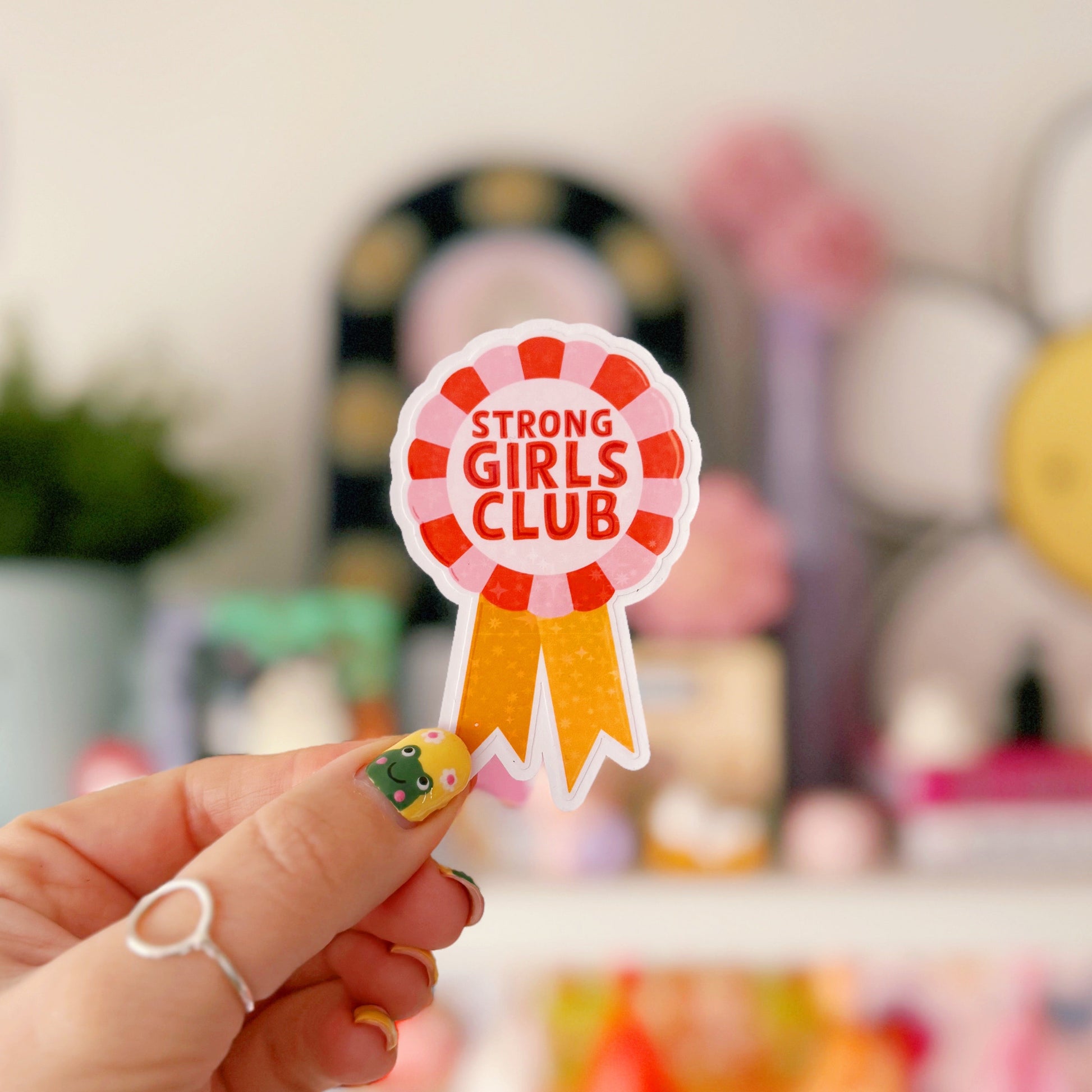A holographic sticker, with the words 'strong girls club' written in the centre of a rosette award, in pink red and yellow colours.