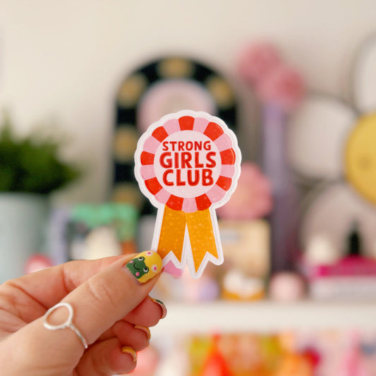 A holographic sticker, with the words 'strong girls club' written in the centre of a rosette award, in pink red and yellow colours.