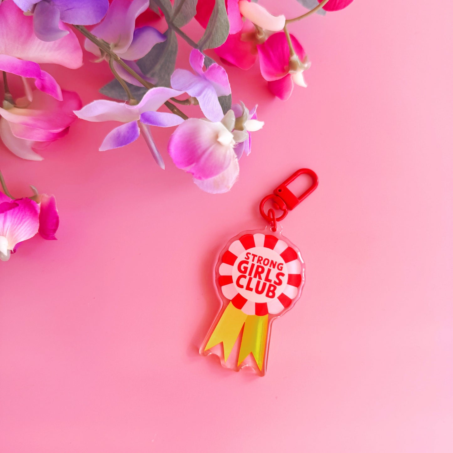 An image showing a key ring or bag charm, with a rosette design. Inside the rosette are the words 'strong girls club'. The colours are pink, red and yellow. 
