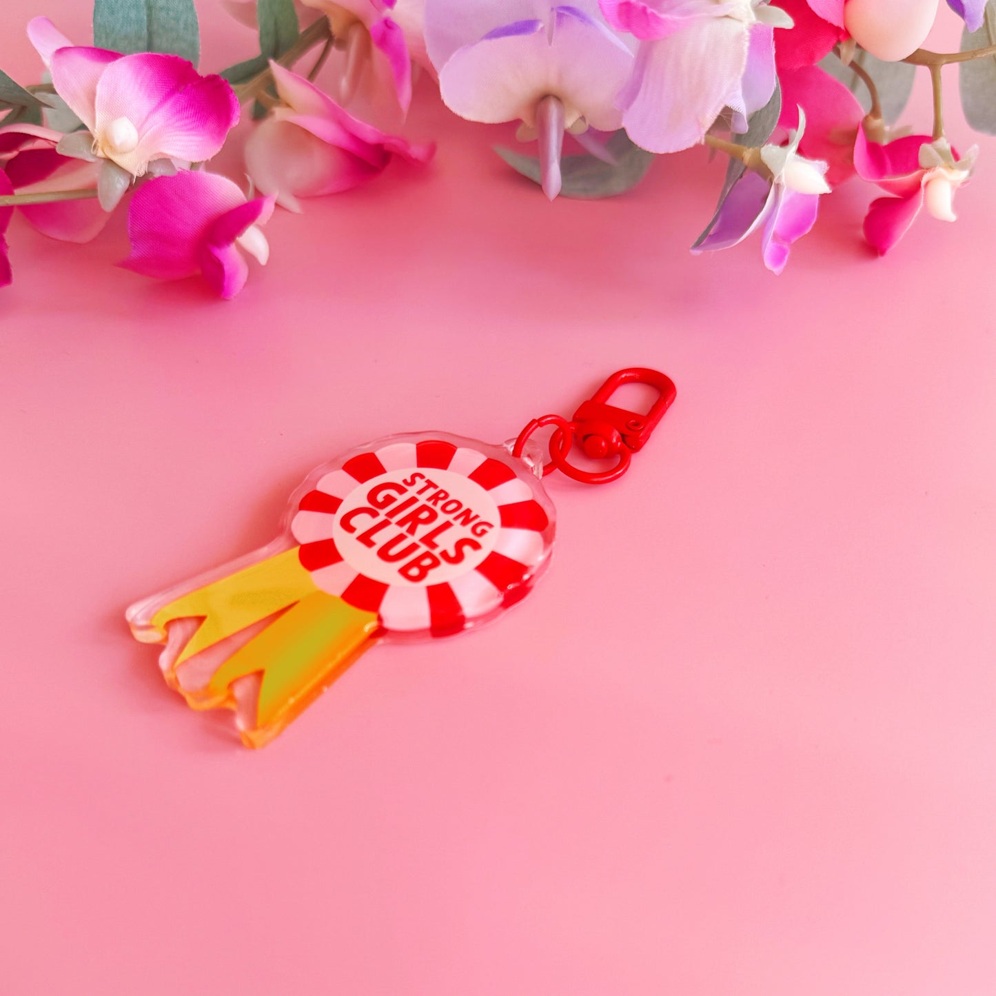 An image showing a key ring or bag charm, with a rosette design. Inside the rosette are the words 'strong girls club'. The colours are pink, red and yellow. 