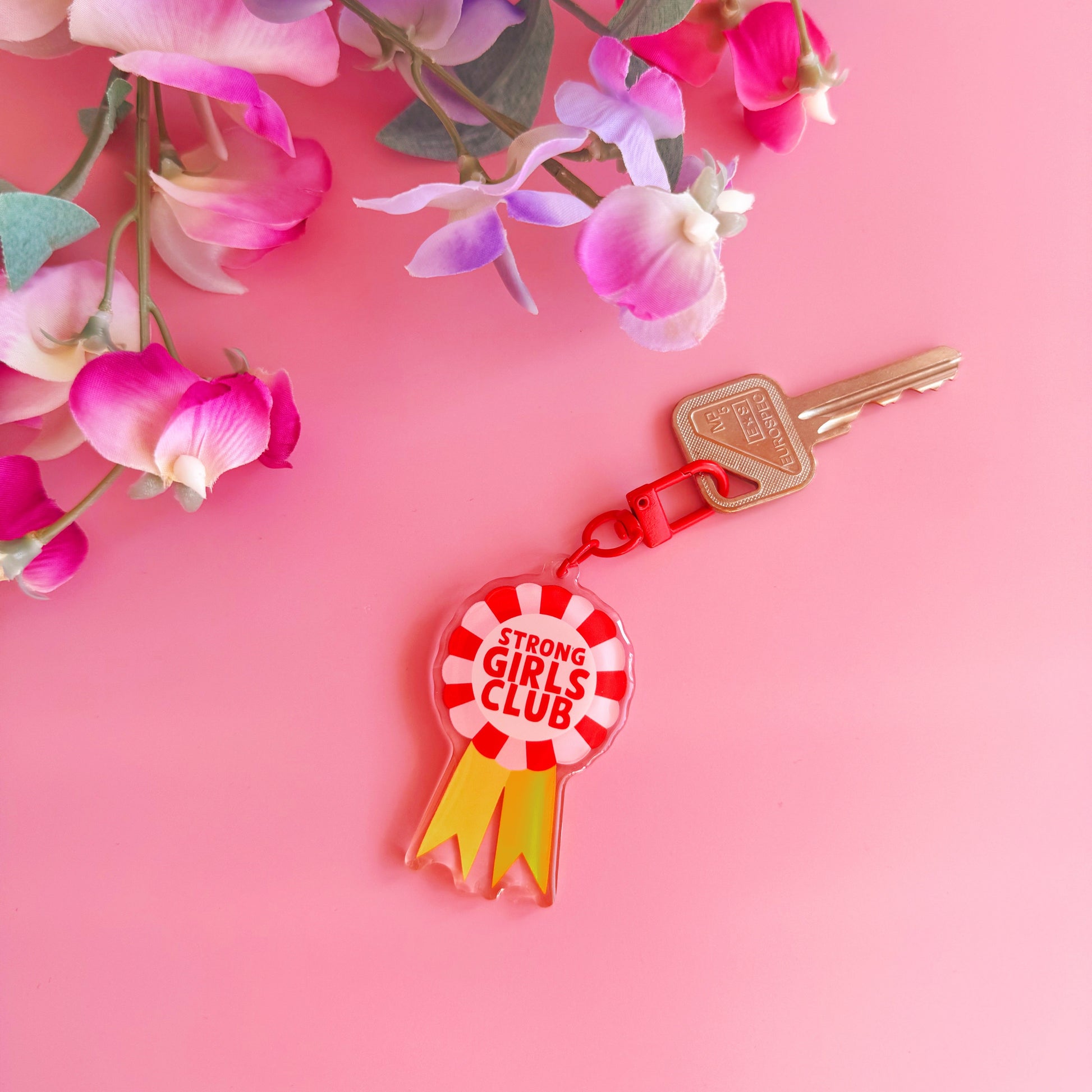 An image showing a key ring or bag charm, with a rosette design. Inside the rosette are the words 'strong girls club'. The colours are pink, red and yellow. 