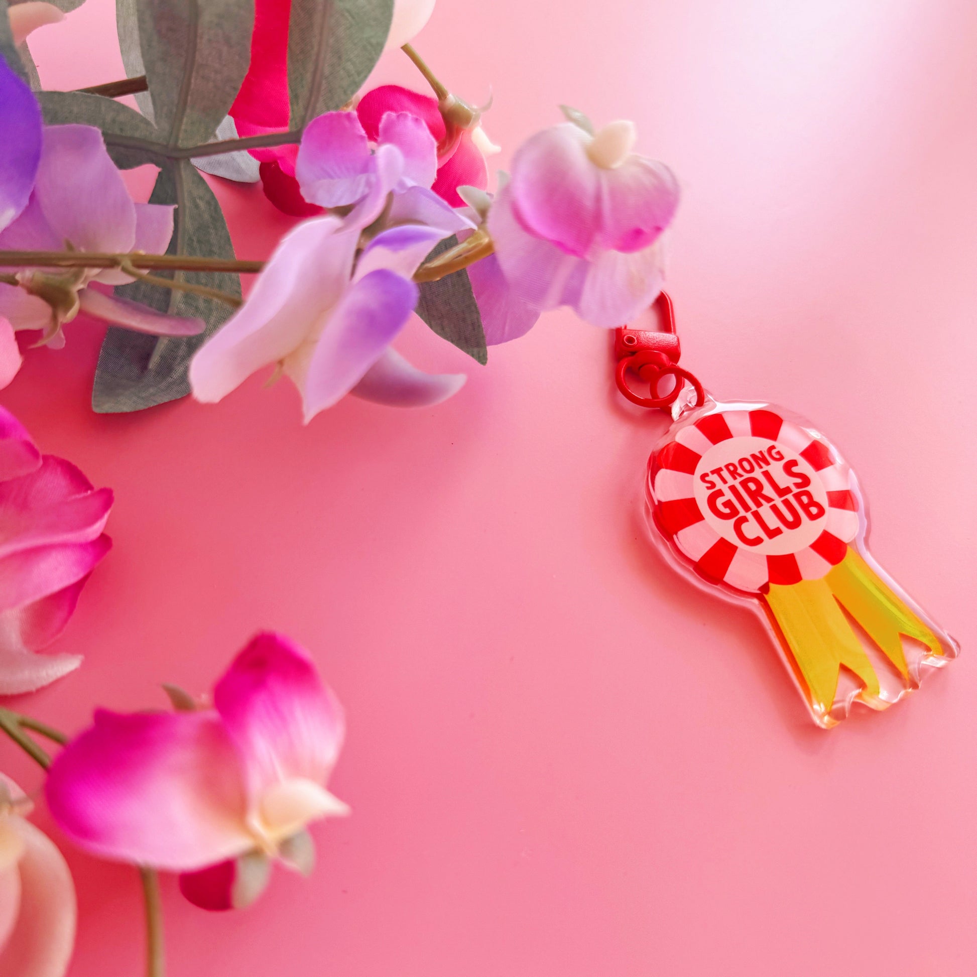 An image showing a key ring or bag charm, with a rosette design. Inside the rosette are the words 'strong girls club'. The colours are pink, red and yellow. 