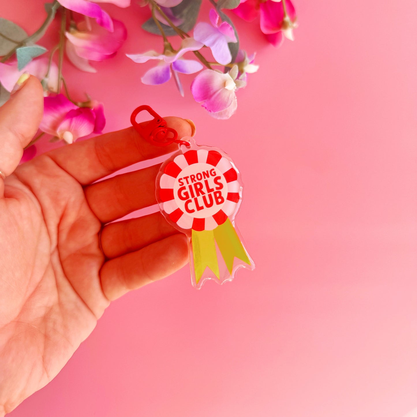 An image showing a key ring or bag charm, with a rosette design. Inside the rosette are the words 'strong girls club'. The colours are pink, red and yellow. 