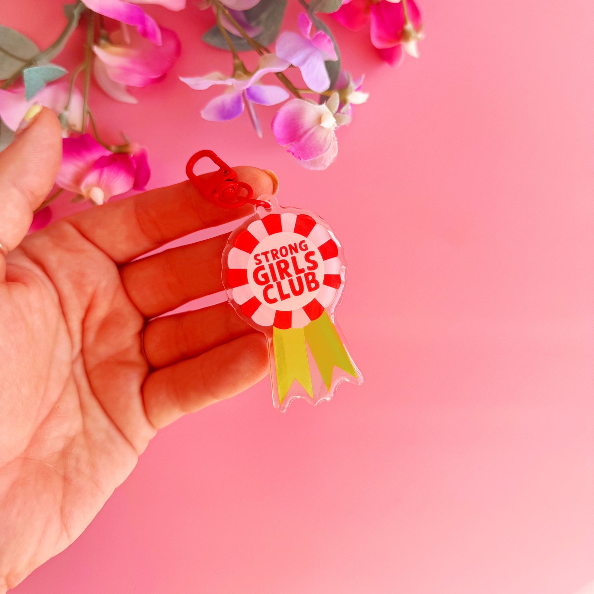 An image showing a key ring or bag charm, with a rosette design. Inside the rosette are the words 'strong girls club'. The colours are pink, red and yellow. 