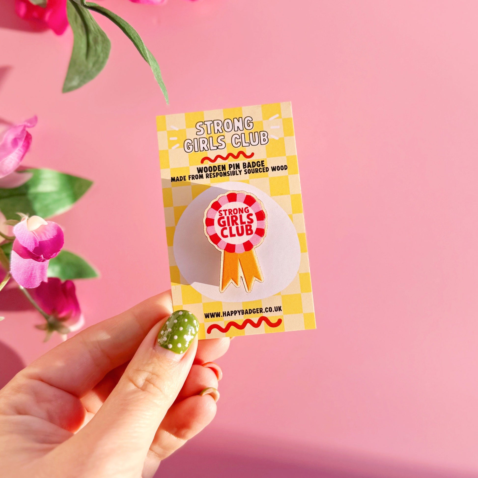 Hand holding a 'Strong Girls Club' badge against a pink background with flowers.