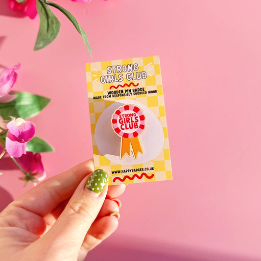 Hand holding a 'Strong Girls Club' badge against a pink background with flowers.