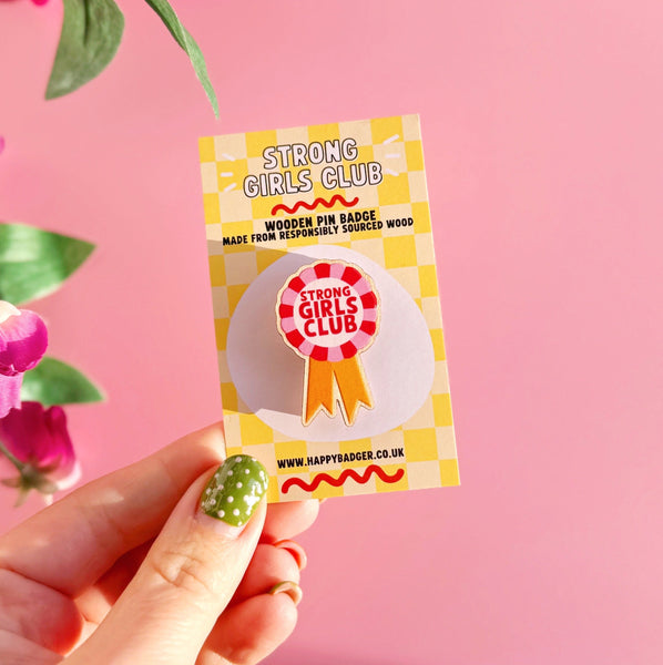 Hand holding a 'Strong Girls Club' badge against a pink background with flowers.