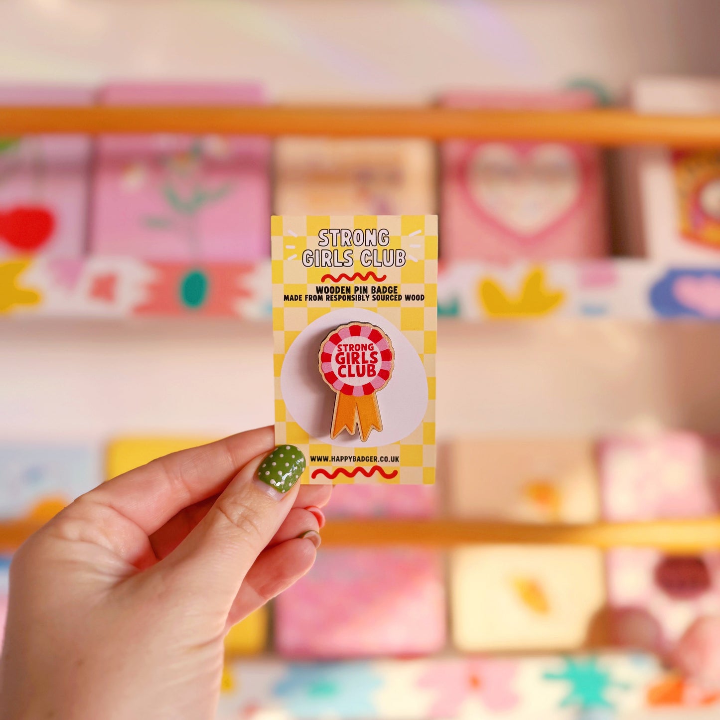 A wooden pin badge, in the shape of a rosette, that says 'strong girls club' in the centre. The colours are red, pink and golden yellow.