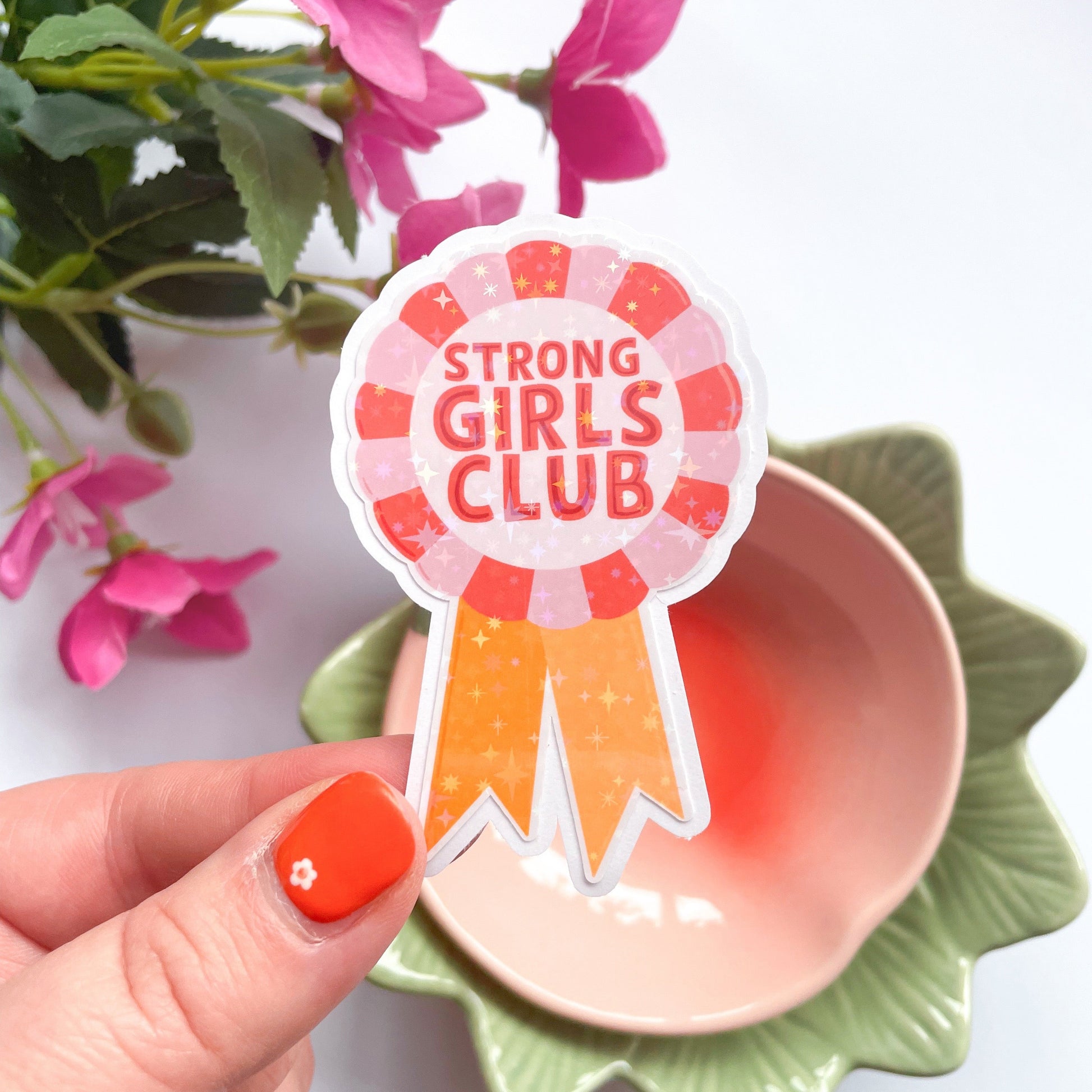 A holographic sticker, with the words 'strong girls club' written in the centre of a rosette award, in pink red and yellow colours.