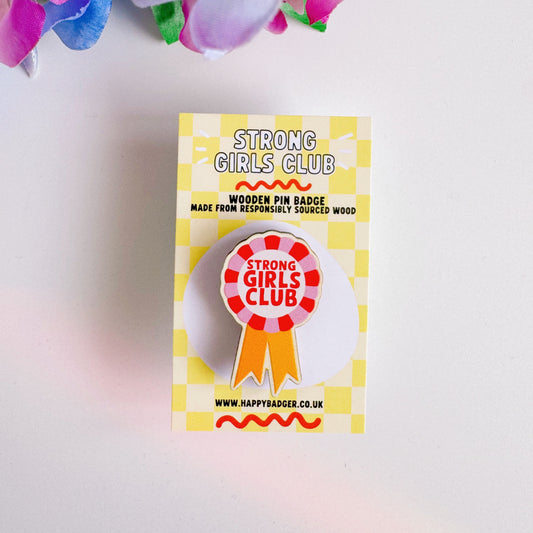 A wooden pin badge with a rosette in pink, red and yellow. The rosette has the words 'strong girls club' in the centre.