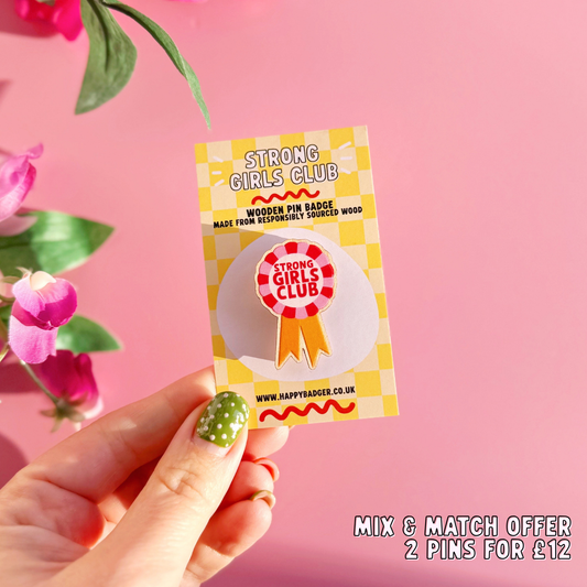 A wooden pin badge, in the shape of a rosette, that says 'strong girls club' in the centre. The colours are red, pink and golden yellow.