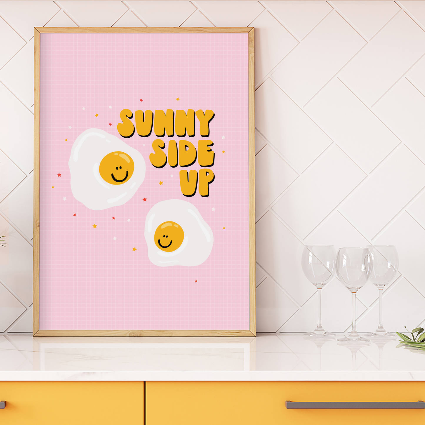 A framed print with a pink background featuring illustrations of sunny side up eggs with smiling faces and the text 'SUNNY SIDE UP' across the top.