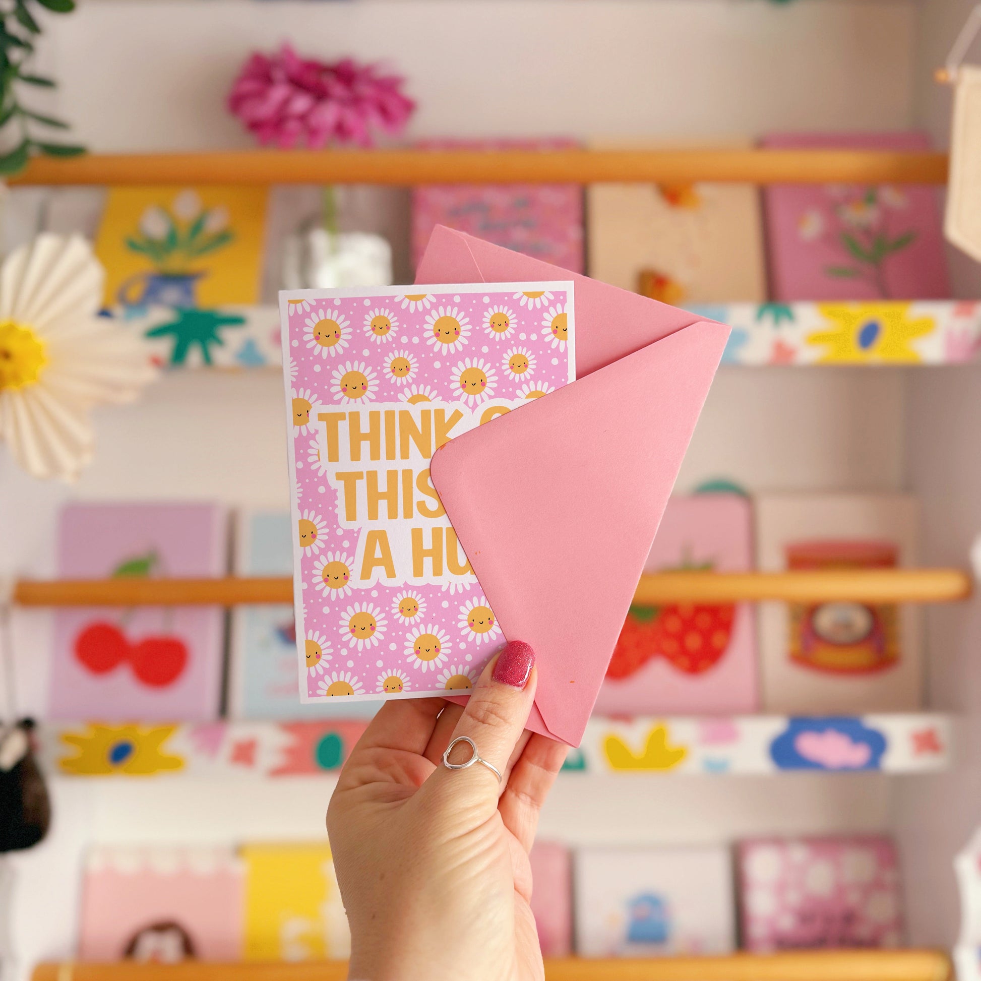 Hand holding a pink card with yellow smiling flowers and text that says 'Think of this as a hug'.