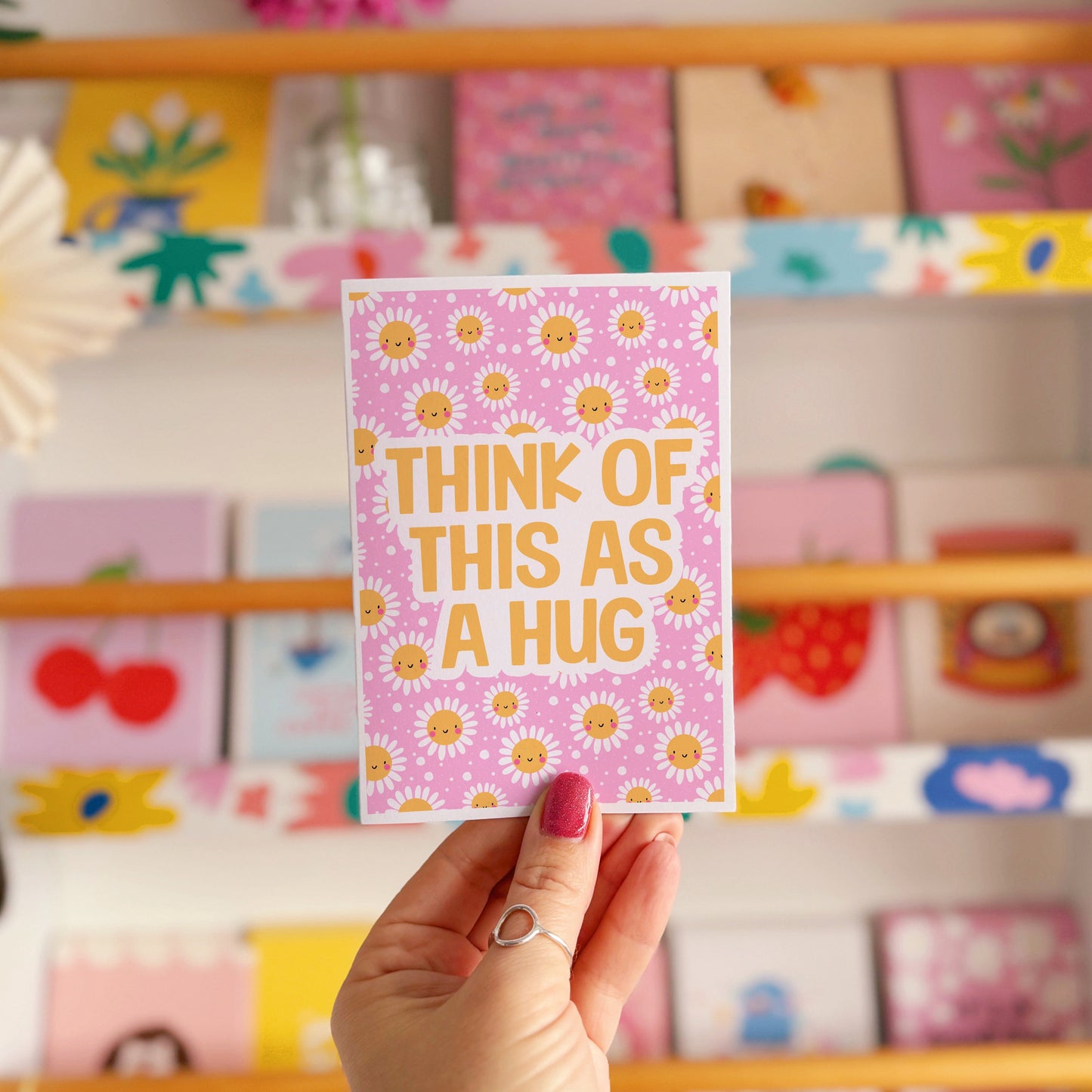 Hand holding a pink card with yellow smiling flowers and text that says 'Think of this as a hug'.