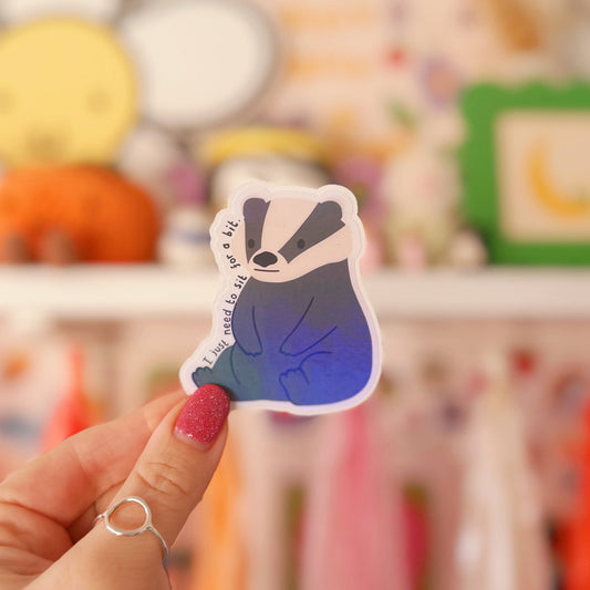 A holographic badger sticker, sitting on its hindquarters, with text around it saying 'I just need to sit for a bit'.