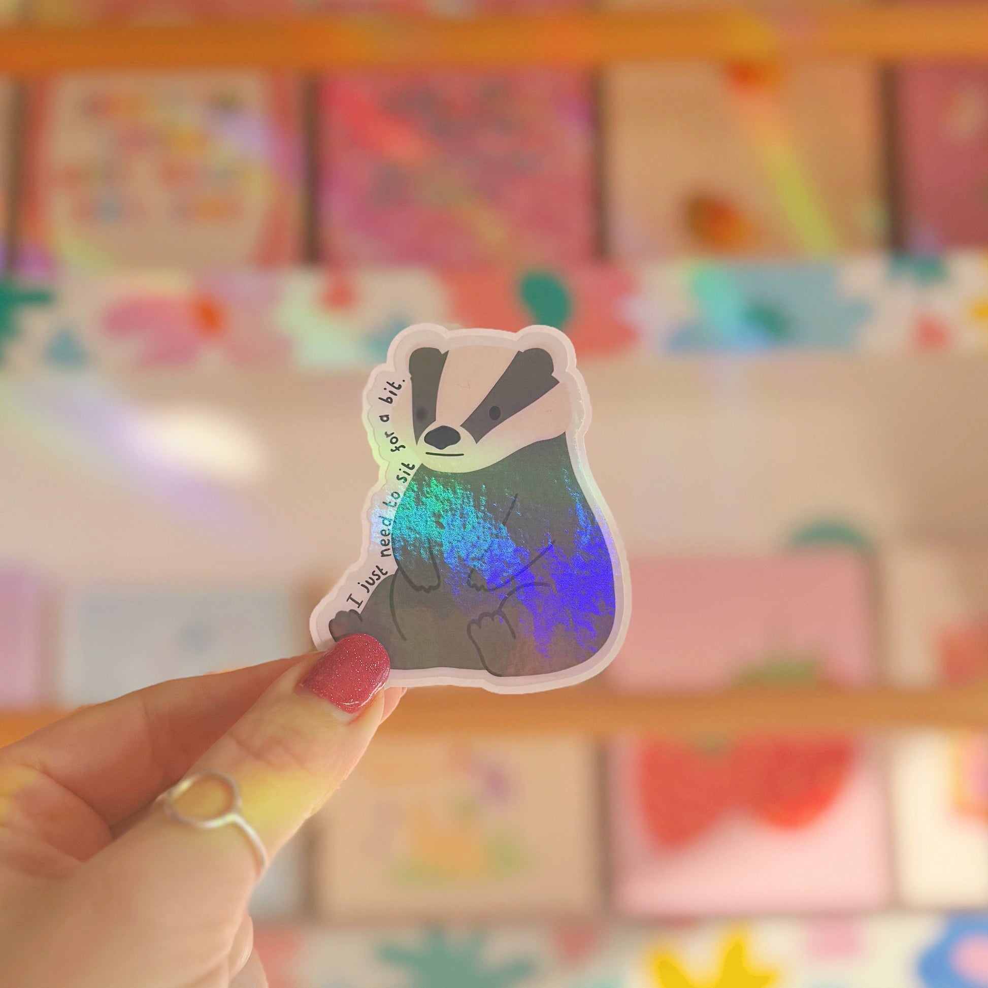 A holographic badger sticker, sitting on its hindquarters, with text around it saying 'I just need to sit for a bit'.
