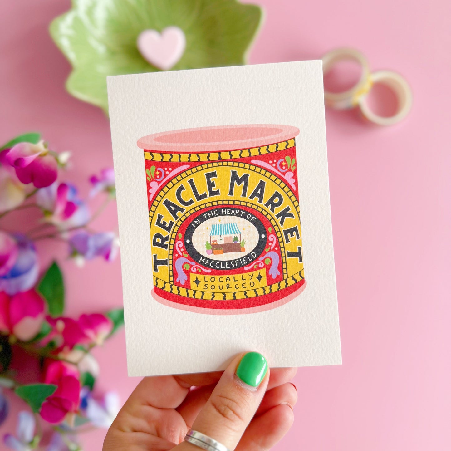 Colourful Treacle Market tin design on a card held by a hand with green nail polish against a pink background.